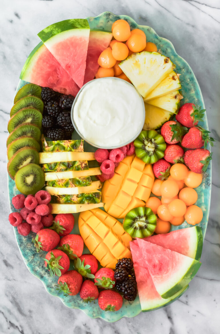A platter with sliced watermelon, pineapple, mango, kiwi, strawberries, raspberries, blackberries, cantaloupe balls, and a bowl of white dip in the center, arranged on a green oval plate.