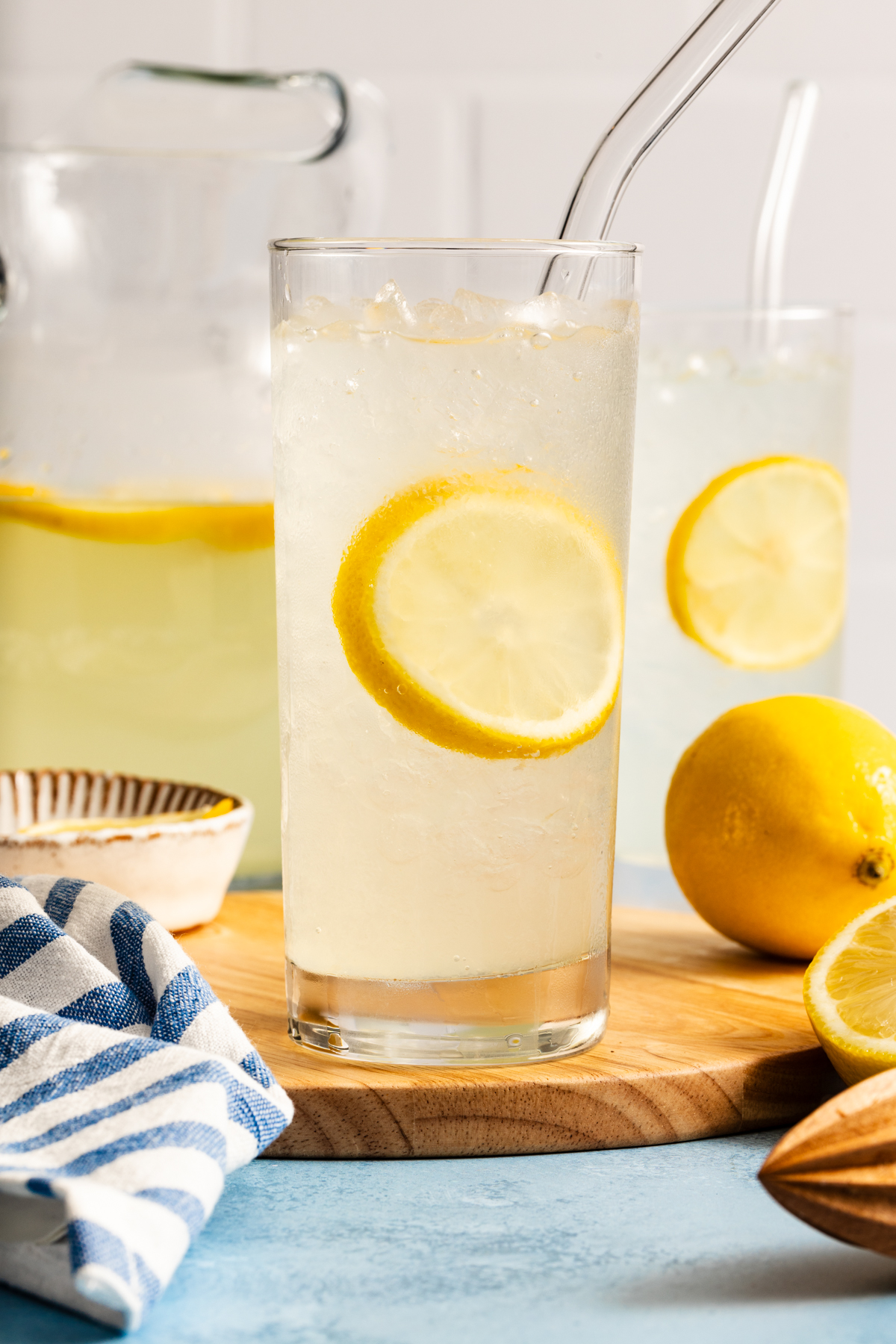 A glass of iced lemonade with a lemon slice and a straw sits on a wooden board. A lemon, pitcher, and another glass of lemonade are in the background, with a blue-striped towel nearby.