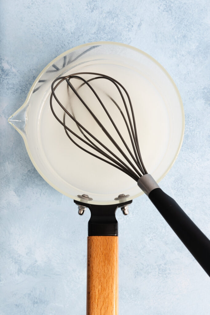 A glass saucepan with a wooden handle, filled with a white liquid as if demonstrating how to make simple syrup, sits on a light blue surface. A black whisk rests inside the pan, mixing the liquid.