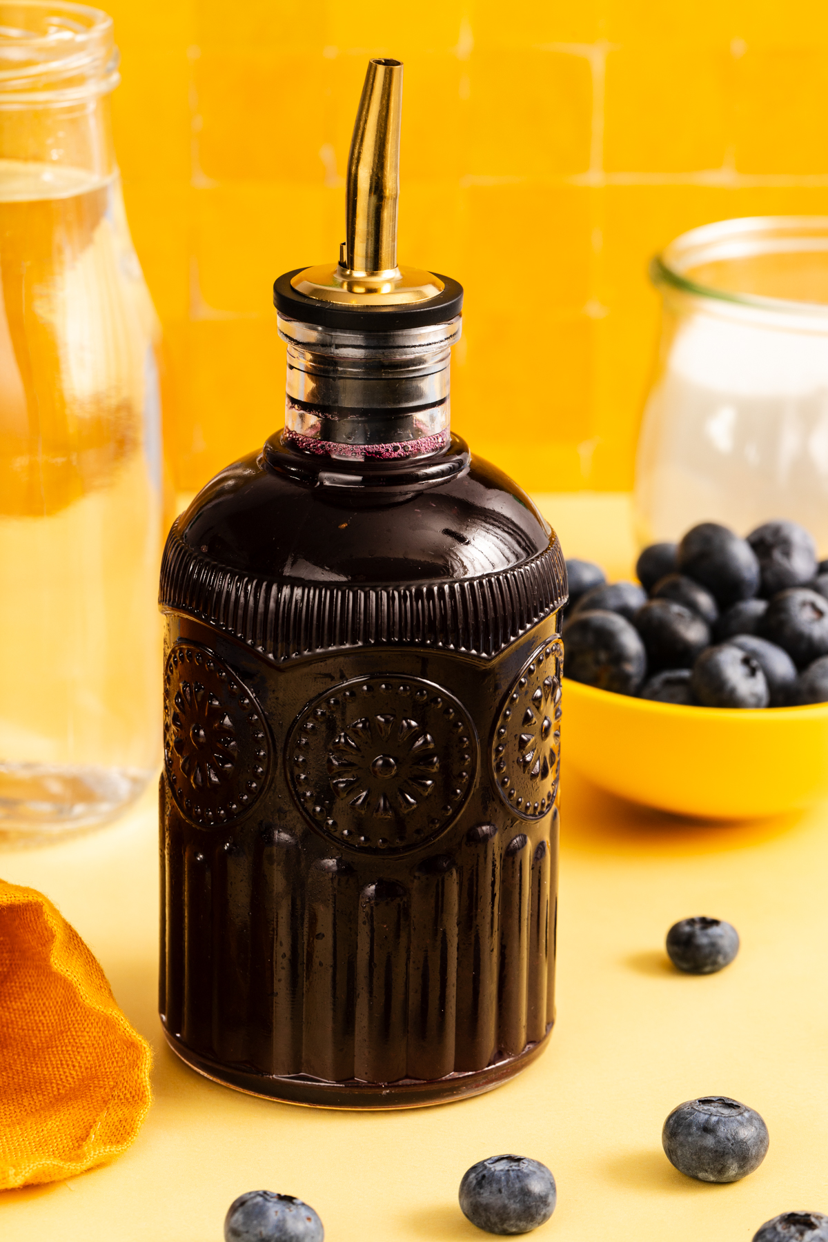 A dark glass bottle with a decorative pattern and metal spout sits on a yellow surface near fresh blueberries, a bowl of blueberries, a glass jar—perfect for storing homemade simple syrup if you’re learning how to make simple syrup—a bottle of water, and an orange napkin, all set against a vibrant yellow tiled background.