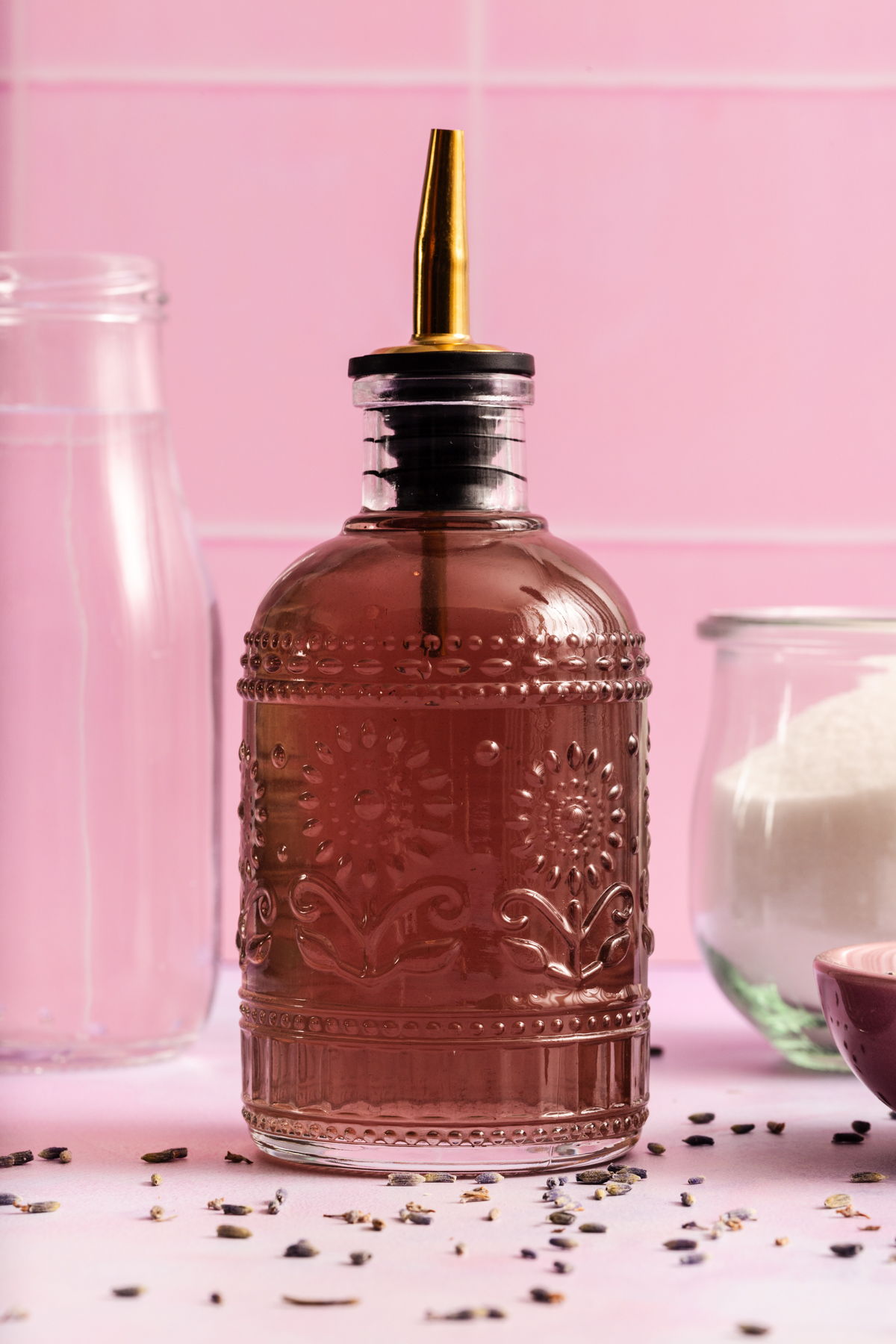 Ornate glass bottle with a golden spout filled with pink liquid, set on a pink surface with scattered dried lavender, a glass of water, and a jar of sugar in the background.
