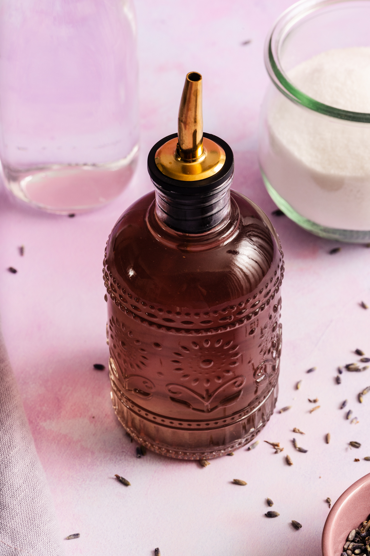 Decorative glass bottle with a metal spout, filled with a brown liquid, sits on a pink surface near a glass jar of sugar and scattered lavender buds.