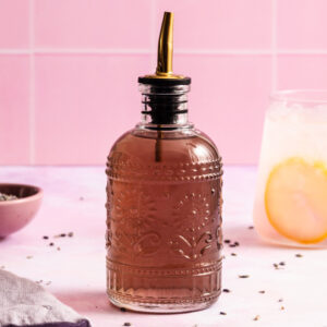 A decorative glass bottle with a metal pour spout is filled with a brown liquid. It sits on a pink surface with a lemon drink and a bowl of lavender in the background.