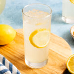 A glass of iced lemonade with a lemon slice inside sits on a wooden board, with a whole lemon and striped cloth nearby.
