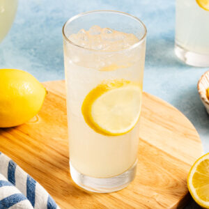 A glass of iced lemonade with a lemon slice inside sits on a wooden board, with a whole lemon and striped cloth nearby.