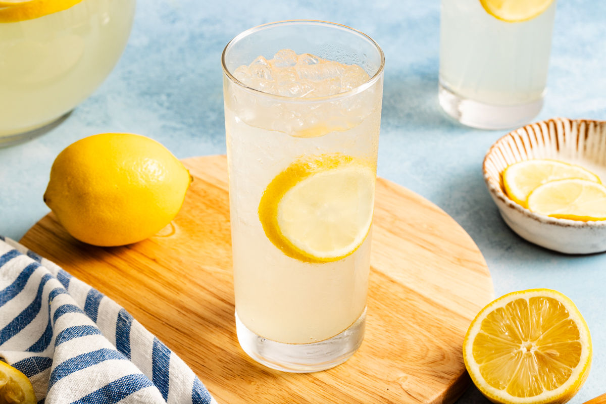 A glass of iced lemonade with a lemon slice sits on a wooden board, surrounded by lemons and lemon slices, on a blue surface with a striped cloth nearby.