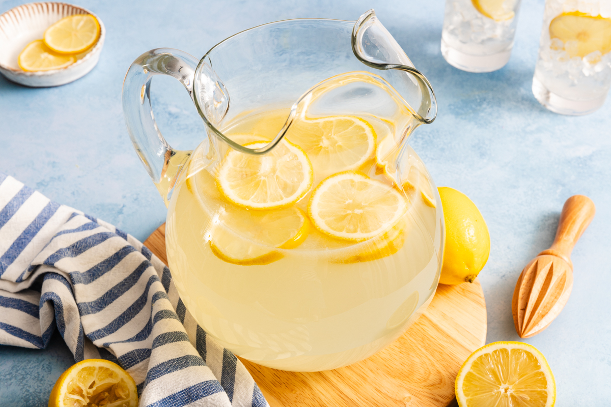 A glass pitcher filled with lemonade and lemon slices sits on a wooden board. A striped towel, a lemon squeezer, and lemon halves are nearby. Two glasses of lemonade with ice and lemon slices are in the background.