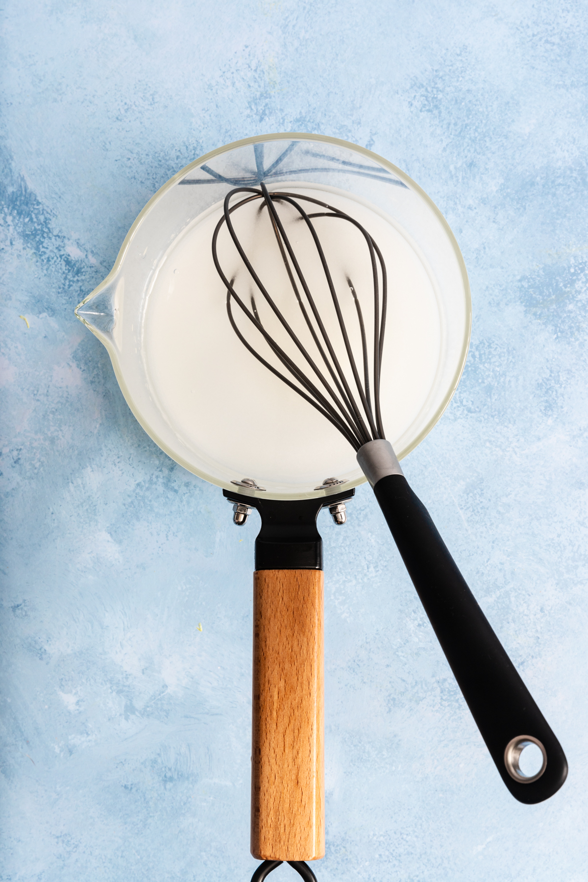 A glass saucepan with a wooden handle, filled with white liquid, sits on a blue surface. A black-handled whisk rests inside the saucepan.