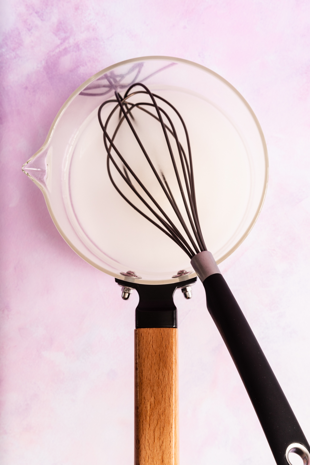 A glass mixing bowl filled with a white liquid sits on a pink surface. A black whisk with a wooden handle rests inside the bowl.