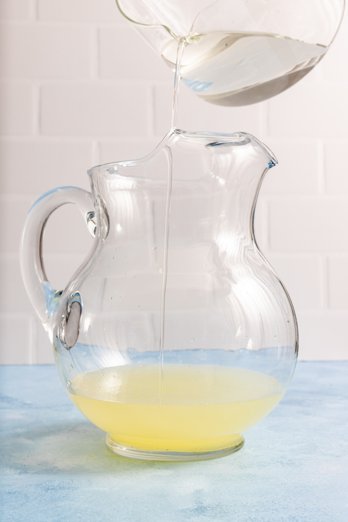 A clear liquid is being poured from a bowl into a glass pitcher containing a small amount of yellow liquid, set on a blue surface with a white tiled background.