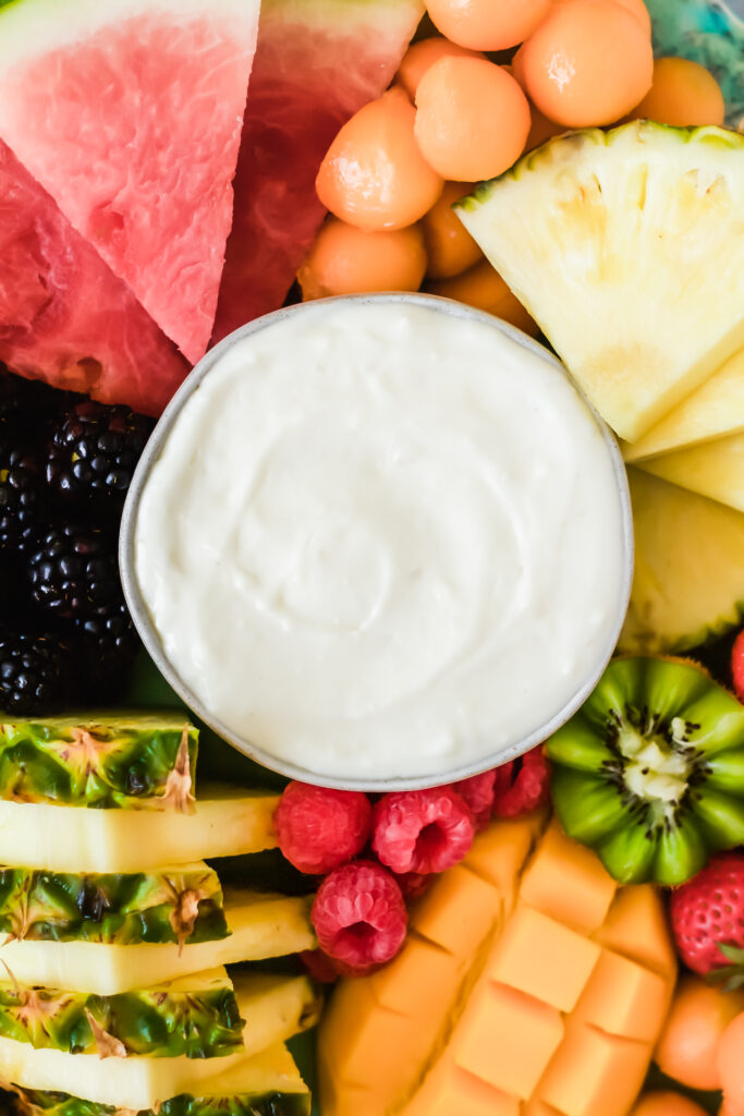 A bowl of creamy white dip surrounded by assorted fresh fruits including watermelon, pineapple, cantaloupe, kiwi, blackberries, raspberries, and mango, arranged closely together.