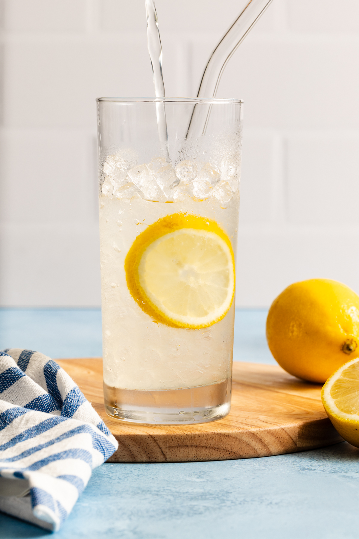 A glass of iced lemon water with a lemon slice and two straws sits on a wooden board, next to a whole lemon, a lemon half, and a striped cloth on a blue surface. Liquid is being poured into the glass.