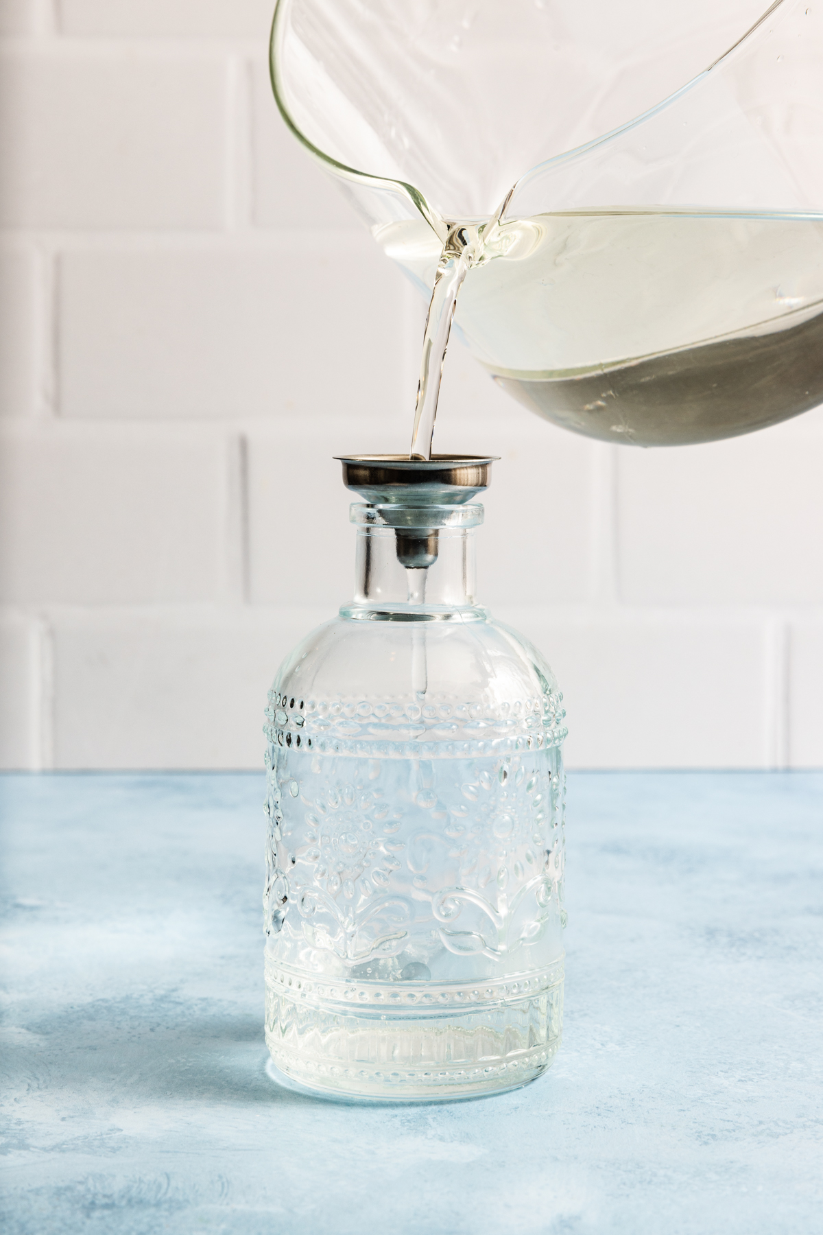 Water is being poured from a glass container through a metal funnel into a decorative clear glass bottle, set against a blue surface and white tiled background—demonstrating an elegant step you might see when learning how to make simple syrup.