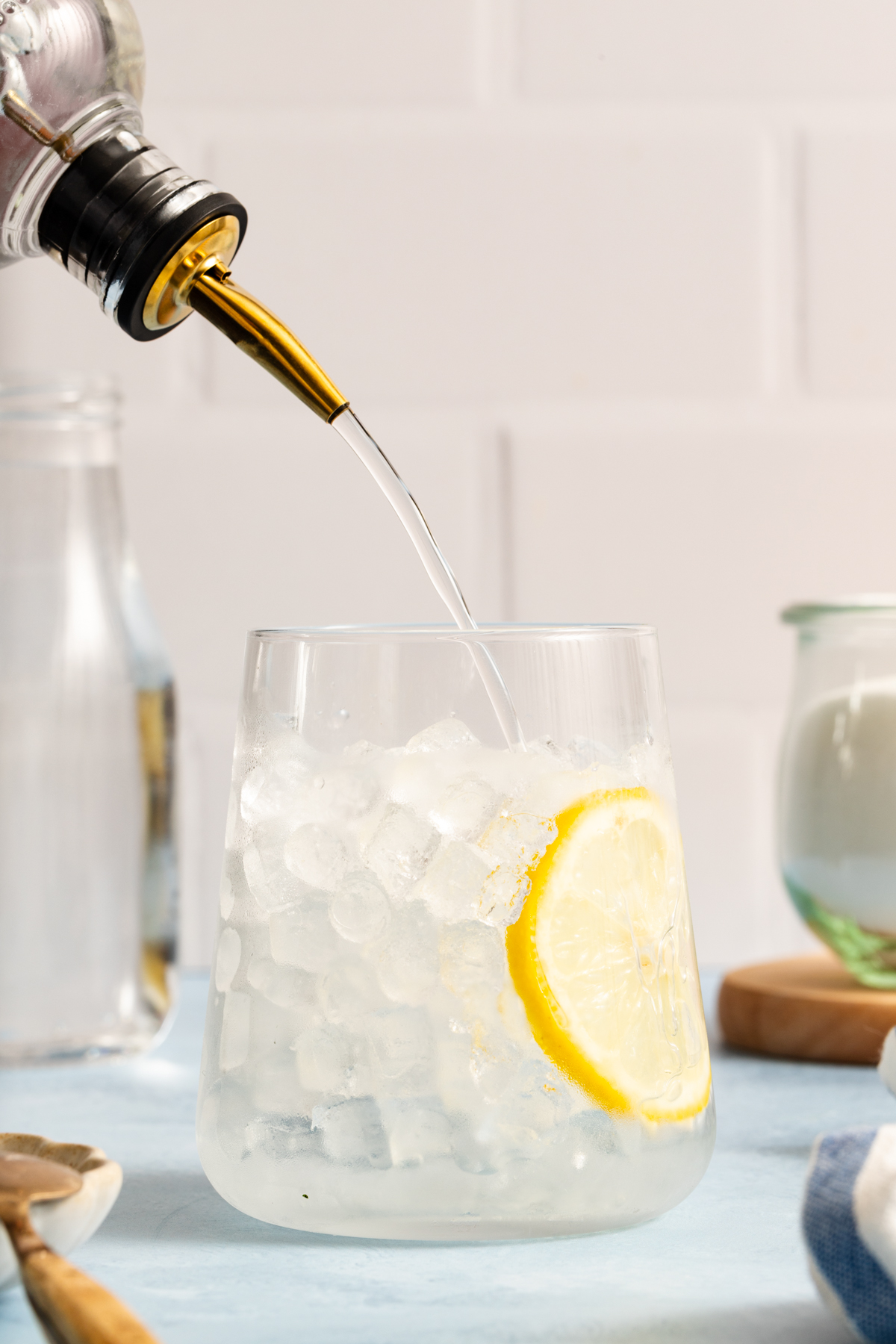 A stream of clear liquid, reminiscent of how to make simple syrup, is being poured into a glass filled with ice and a lemon slice. The scene is set on a light blue surface with blurred kitchen items in the background.