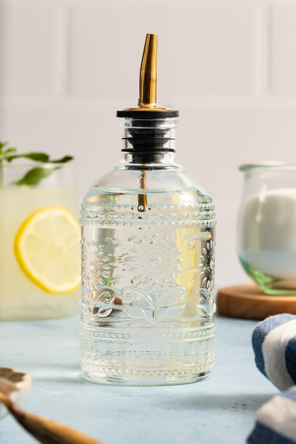A decorative glass bottle with a metal spout sits on a blue surface, ready to showcase how to make simple syrup in style. In the background, there’s a drink with a lemon slice, a glass jar, and a striped cloth adding vibrant touches to the scene.