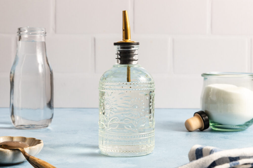 A glass bottle with a spout filled with clear liquid sits on a blue surface—a scene reminiscent of how to make simple syrup. Nearby are a glass jar of sugar, a jar of water, a striped cloth, and a metal spoon.