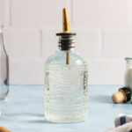 A clear glass oil dispenser with a decorative pattern and a gold spout sits on a light blue countertop against a white tiled wall, evoking the kind of charming setting where you might learn how to make simple syrup. Other kitchen items are partially visible in the background.