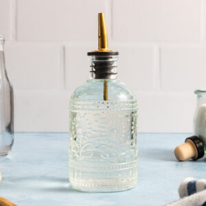 A clear glass oil dispenser with a decorative pattern and a gold spout sits on a light blue countertop against a white tiled wall, evoking the kind of charming setting where you might learn how to make simple syrup. Other kitchen items are partially visible in the background.