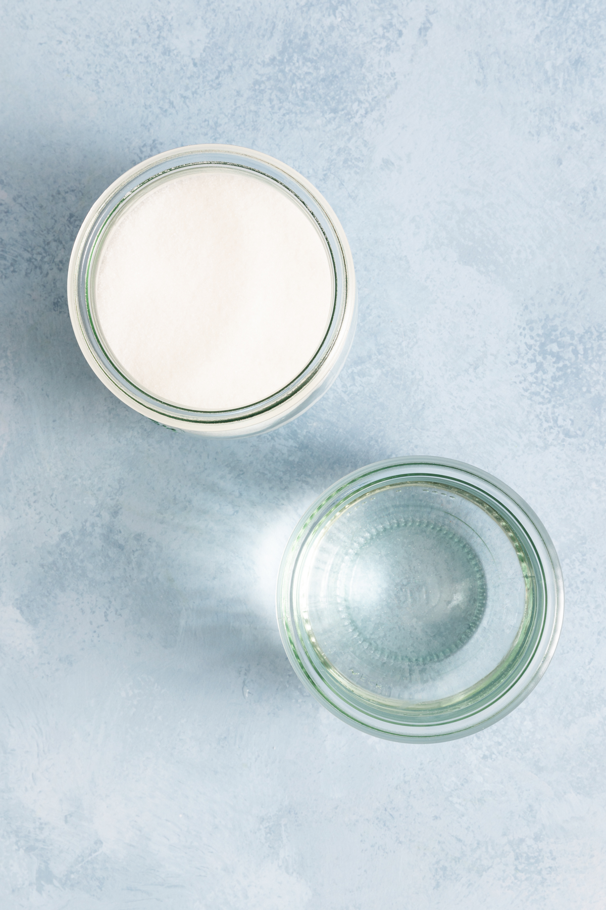 Two clear glass bowls on a light blue surface, one filled with white granulated sugar and the other with clear liquid, likely water, seen from above—ingredients often used when learning how to make simple syrup.