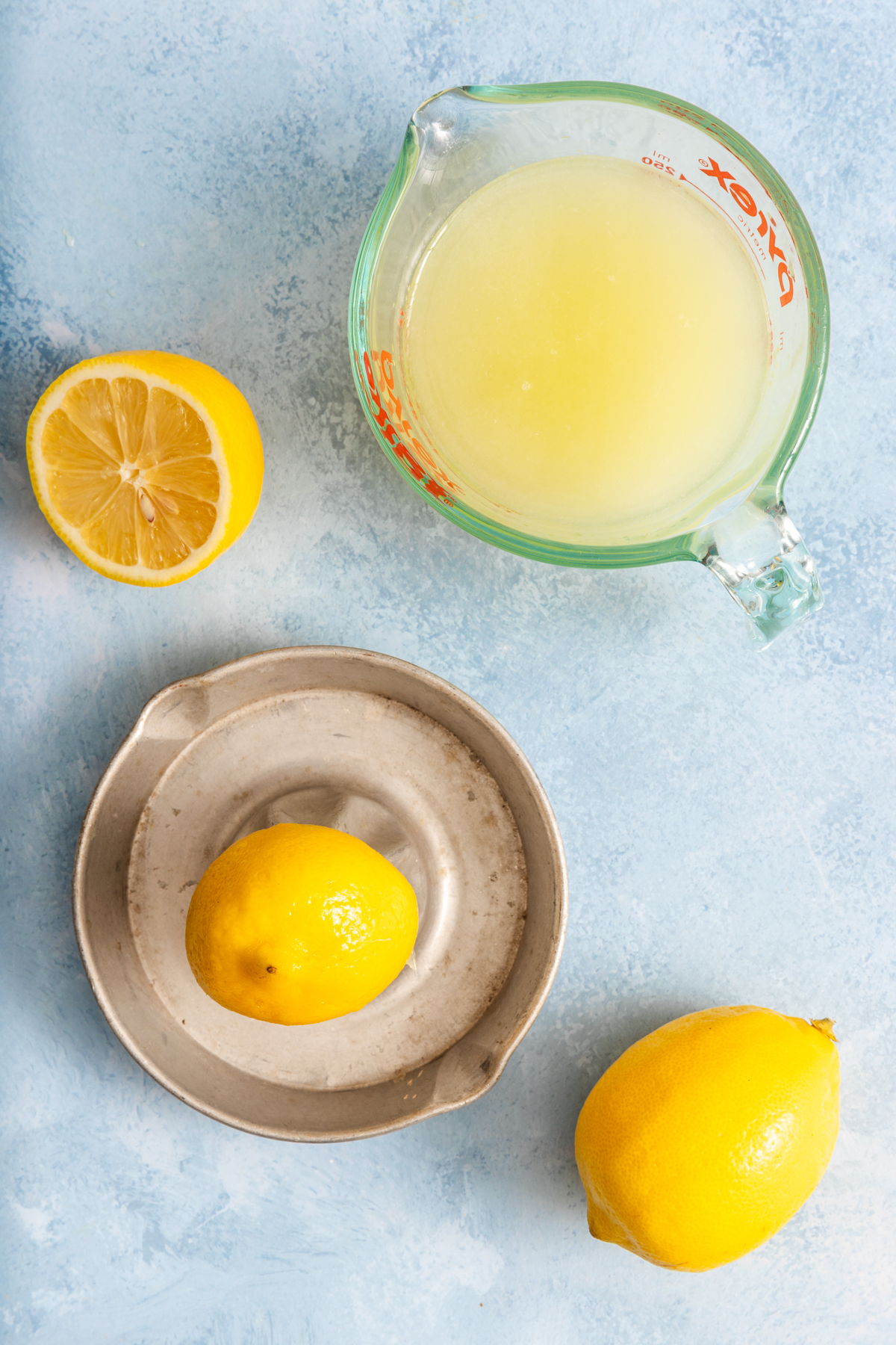 A glass measuring cup of lemon juice, a halved lemon, a whole lemon on a plate, and another whole lemon sit on a light blue surface.