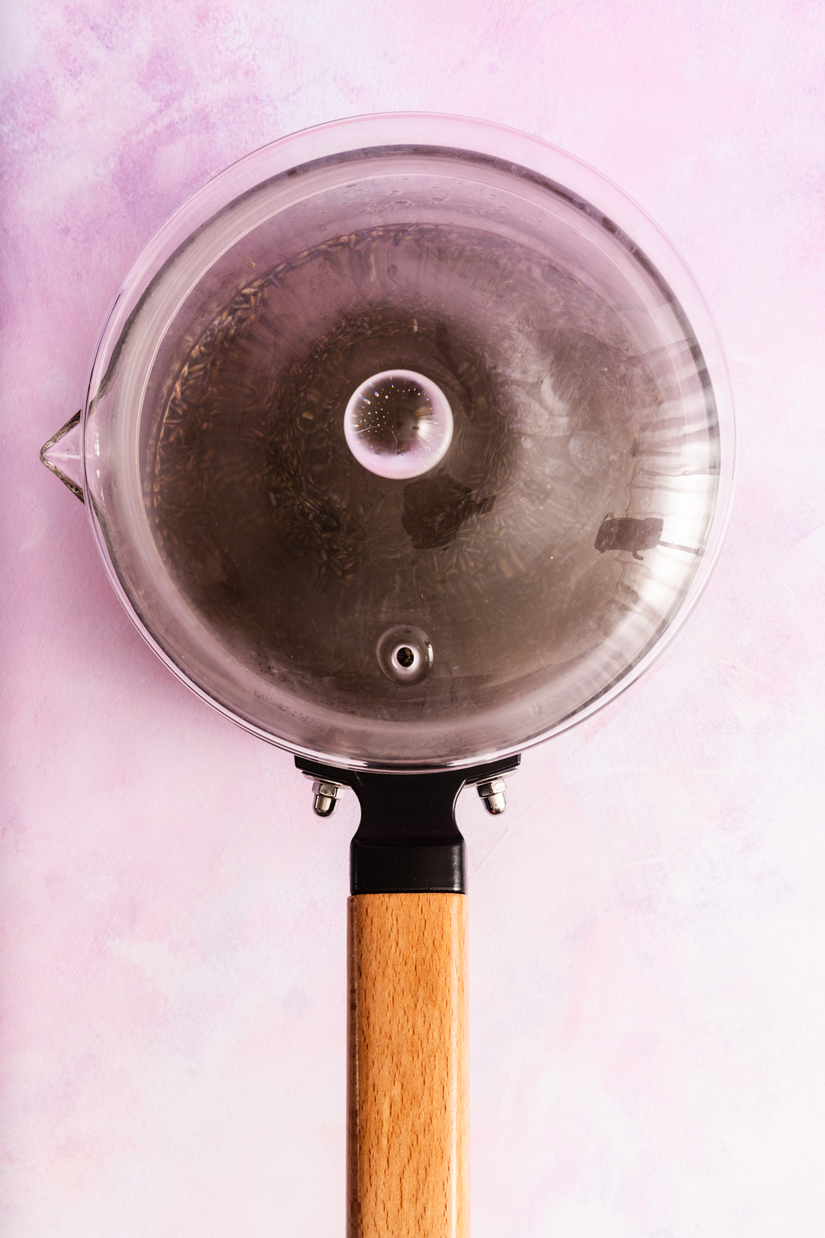 A glass saucepan with a wooden handle, filled with boiling water and herbs, sits on a light pink textured surface, viewed from above.
