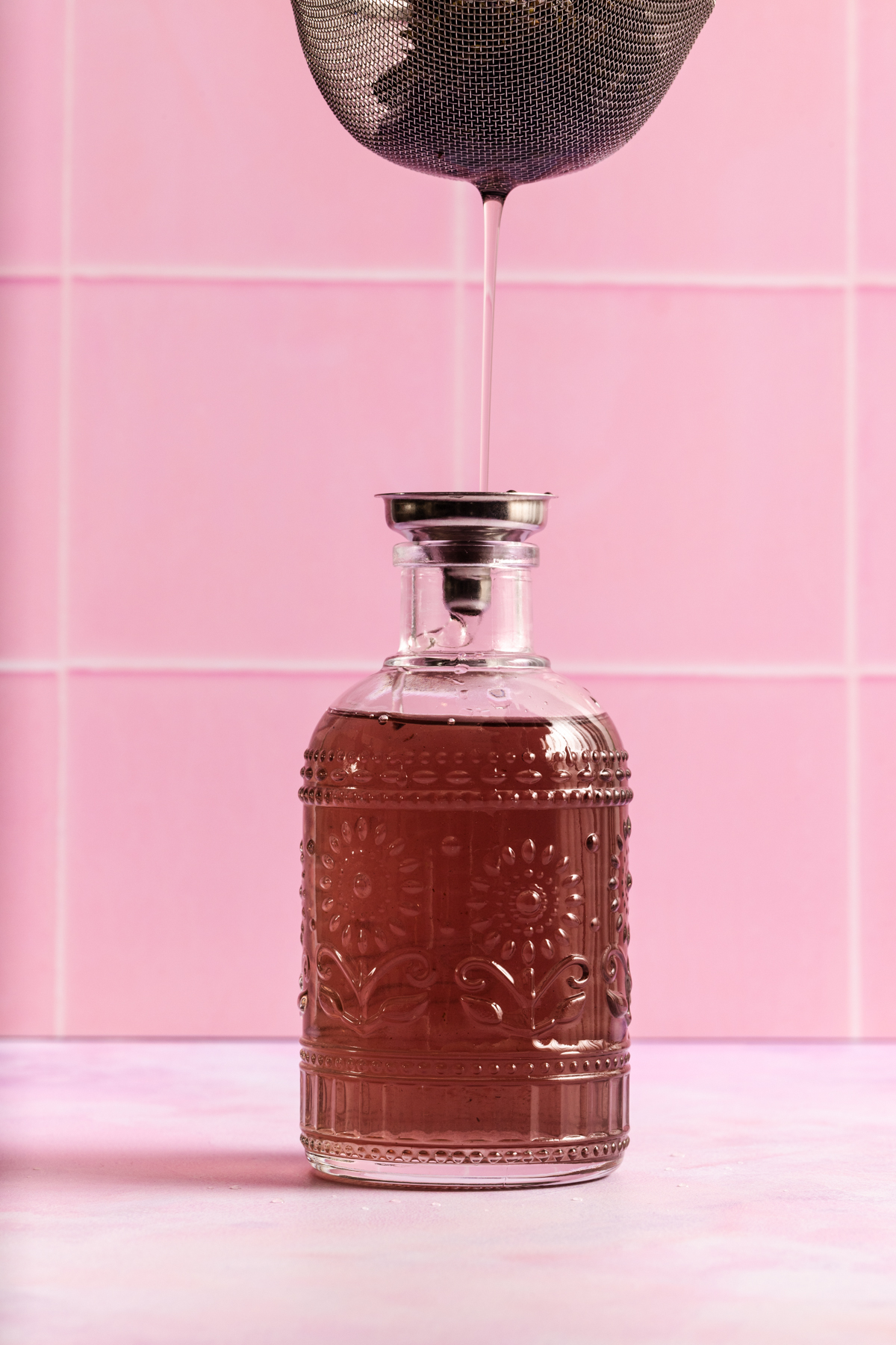 A metal strainer pours liquid into a decorative glass bottle filled with pinkish liquid, set against a pink tiled background.