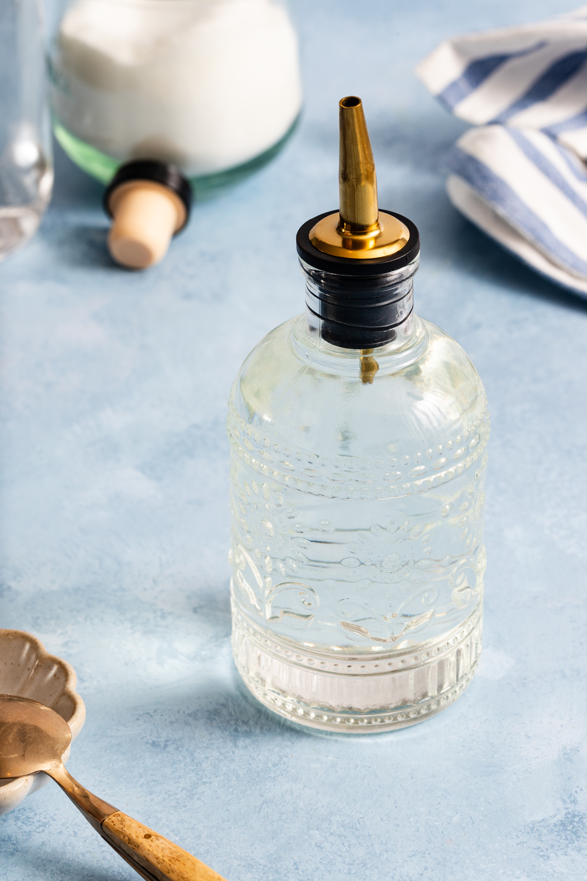 A decorative glass bottle with a gold pour spout sits on a blue surface, surrounded by a spoon, a white dish, a jar of sugar—a perfect setup for learning how to make simple syrup—and a blue-and-white striped cloth.
