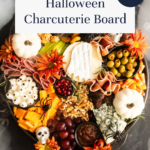 A Halloween-themed charcuterie board with meats, cheeses, olives, grapes, crackers, white pumpkins, orange flowers, candy eyeballs, and plastic spiders. Text says Halloween Charcuterie Board and Fun & Spooky.