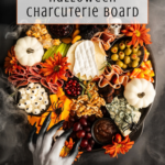 A Halloween charcuterie board with meats, cheeses, fruits, fake spider webs, mini pumpkins, and a spooky gray hand. Text reads: How to make a Halloween Charcuterie Board. Logo: off the eaten path..