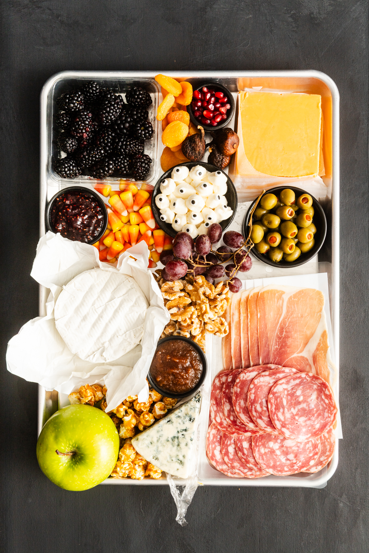 A tray with assorted cheeses, sliced meats, green olives, crackers, blackberries, grapes, dried fruits, walnuts, popcorn, an apple, jam, and pomegranate seeds arranged in separate sections.