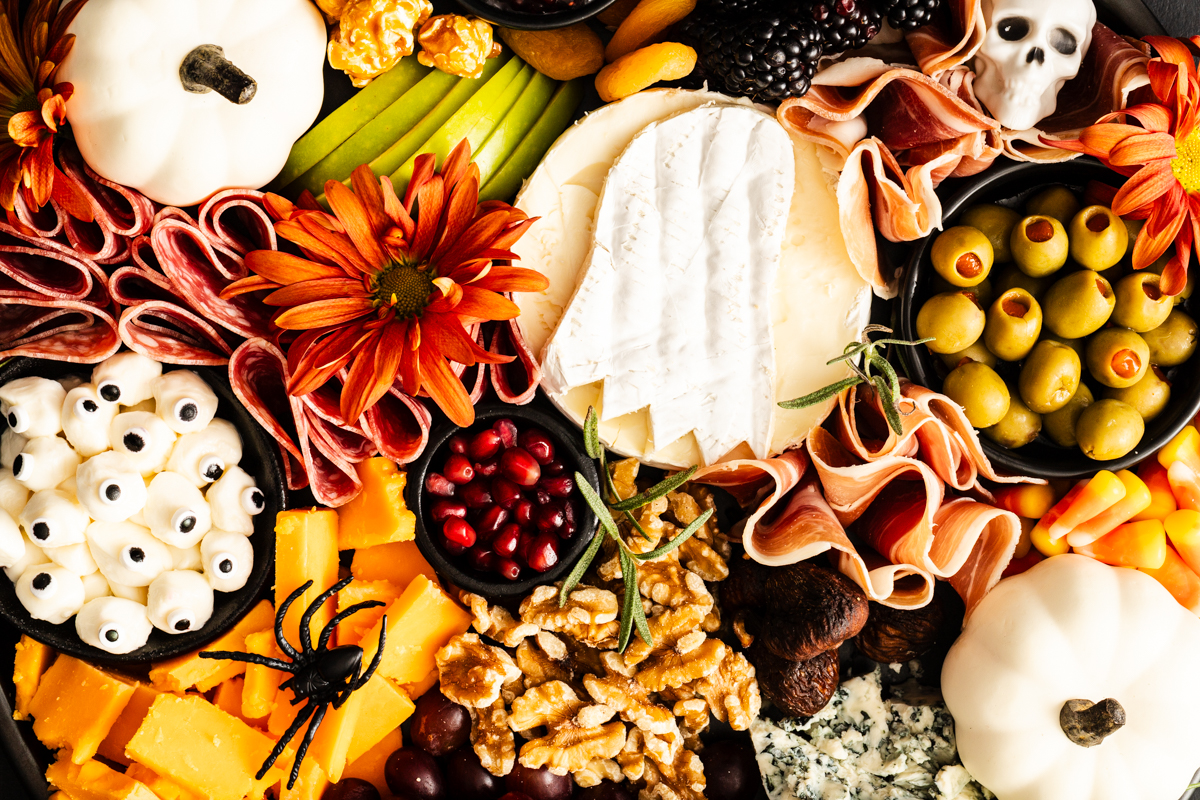 A Halloween-themed charcuterie board with cheeses, meats, olives, fruits, nuts, white pumpkins, candy eyeballs, a plastic spider, flowers, and a white chocolate skull, arranged festively for the holiday.