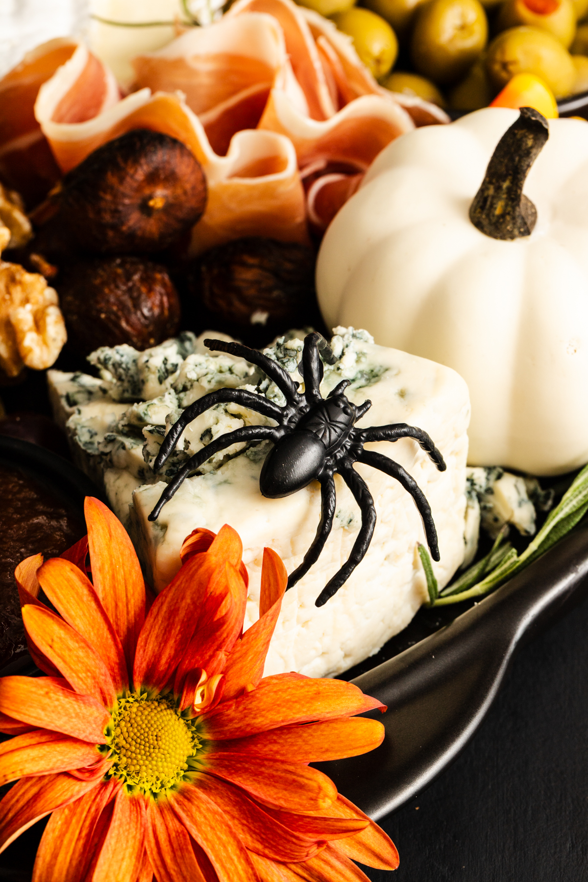 A plastic black spider rests on blue cheese next to an orange flower, a white pumpkin, prosciutto, figs, olives, and walnuts on a charcuterie board.