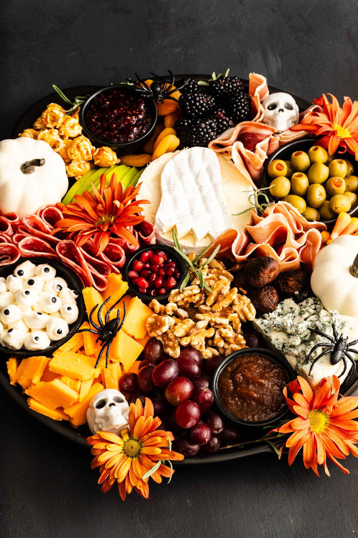 A Halloween-themed charcuterie board with cheeses, meats, grapes, nuts, olives, dip, blackberries, caramel popcorn, candy eyes, small white pumpkins, orange flowers, and decorative fake spiders and skulls.