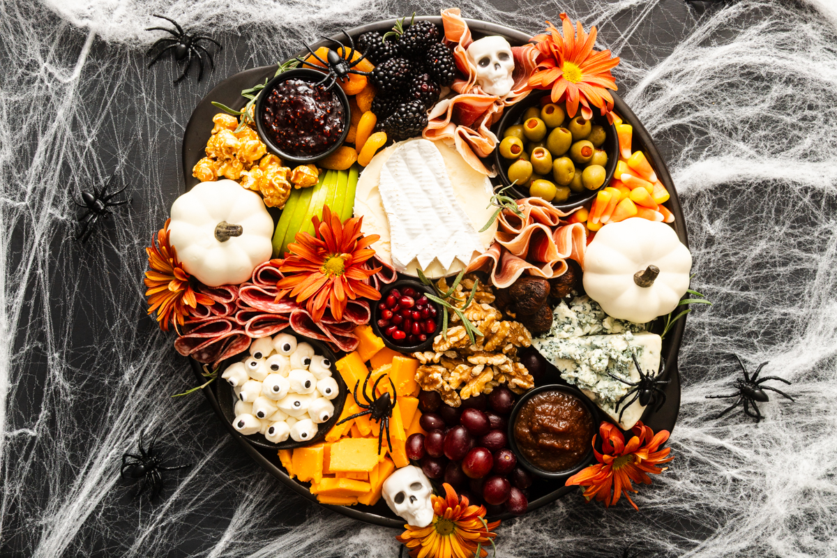Halloween-themed charcuterie board with cheese, meats, olives, fruit, popcorn, candy eyes, mini pumpkins, flowers, and skull decorations on a black platter, surrounded by fake cobwebs and plastic spiders.