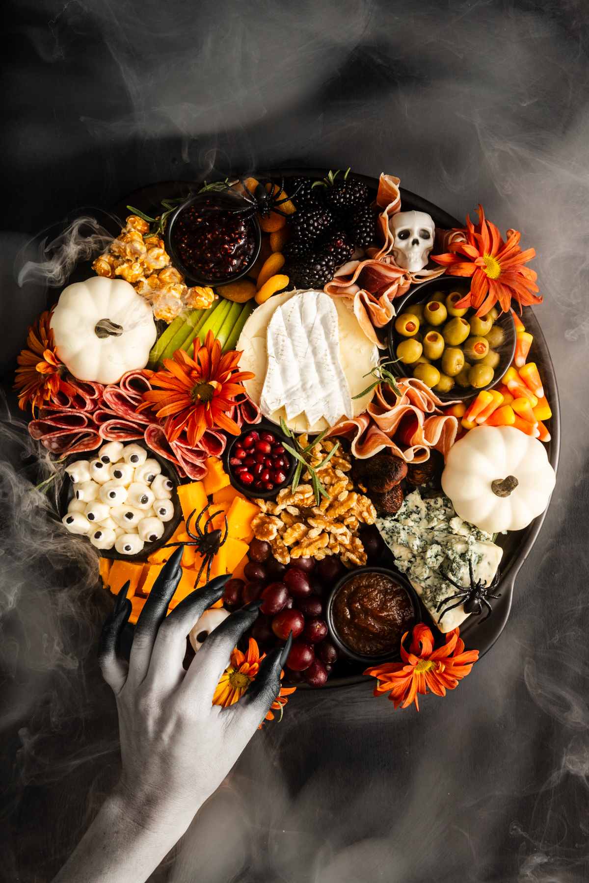 A Halloween-themed charcuterie board with pumpkins, cheeses, fruit, and spooky decorations. A white hand with black nails reaches toward the food, surrounded by fog. Orange flowers accent the arrangement.