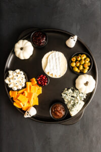 A round black tray with cheeses, olives, sauces, candy eyeballs, pomegranate seeds, white pumpkins, and small skull decorations, arranged for a Halloween-themed snack platter.