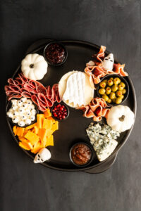 Halloween-themed charcuterie board with cheeses, meats, olives, crackers, small pumpkins, fruit, and dips arranged decoratively on a dark circular tray, including eye-shaped cheese pieces and ghost-shaped decorations.