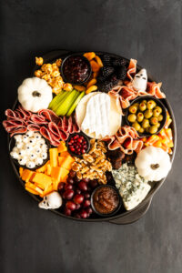 A round platter with cheeses, cured meats, olives, fruits, nuts, and dips, decorated with mini white pumpkins and candy eyes on a dark background for a Halloween-themed charcuterie board.