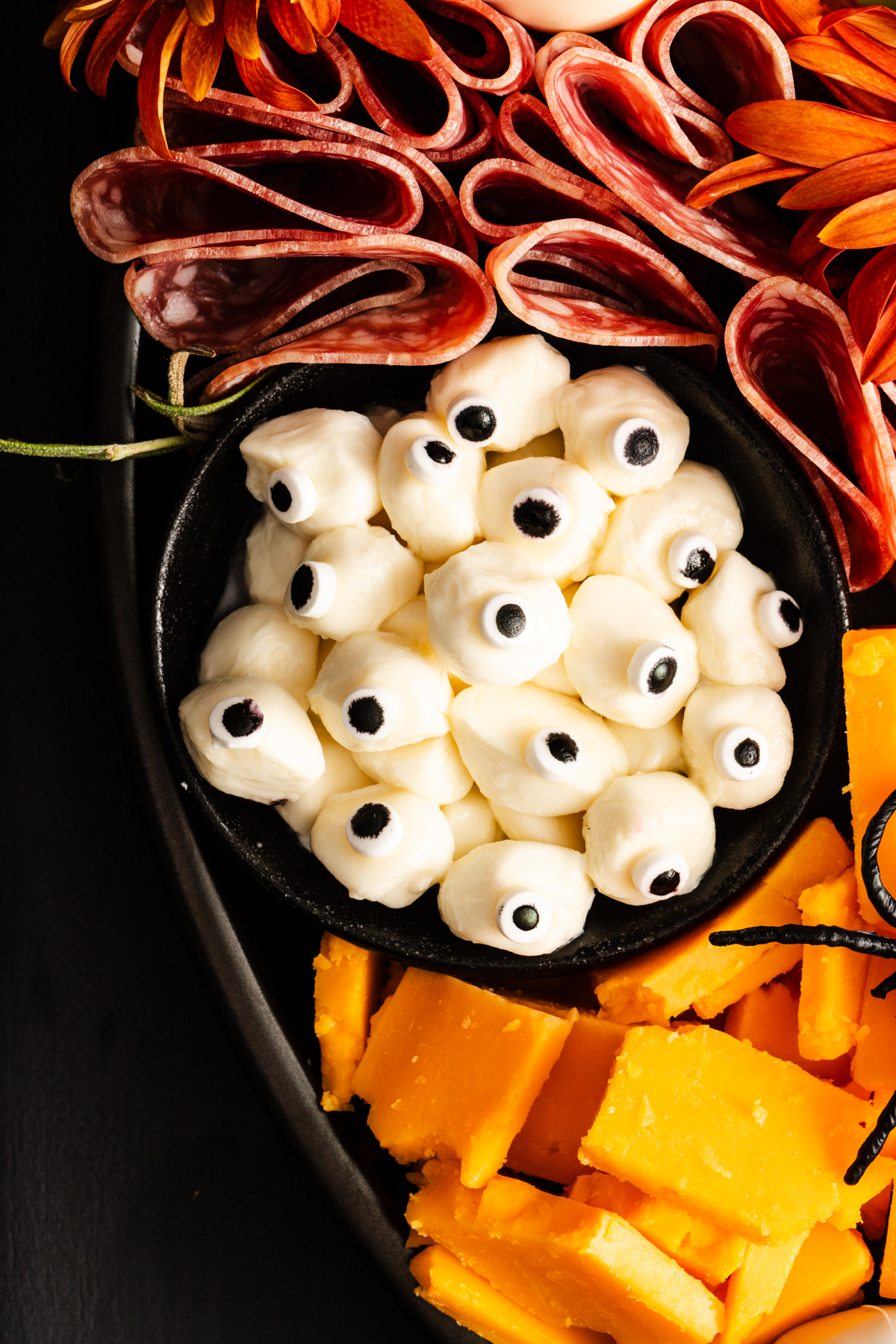 A Halloween charcuterie board with cheese cubes, folded cold cuts, and a bowl of mozzarella balls decorated to look like eyeballs with black olive slices as pupils.