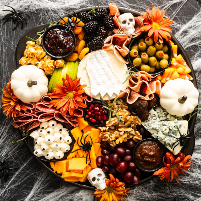 A Halloween-themed charcuterie board with cheese, meats, olives, fruit, popcorn, nuts, flowers, candy eyeballs, white pumpkins, skull decorations, and spider props on a dark background with cobwebs.