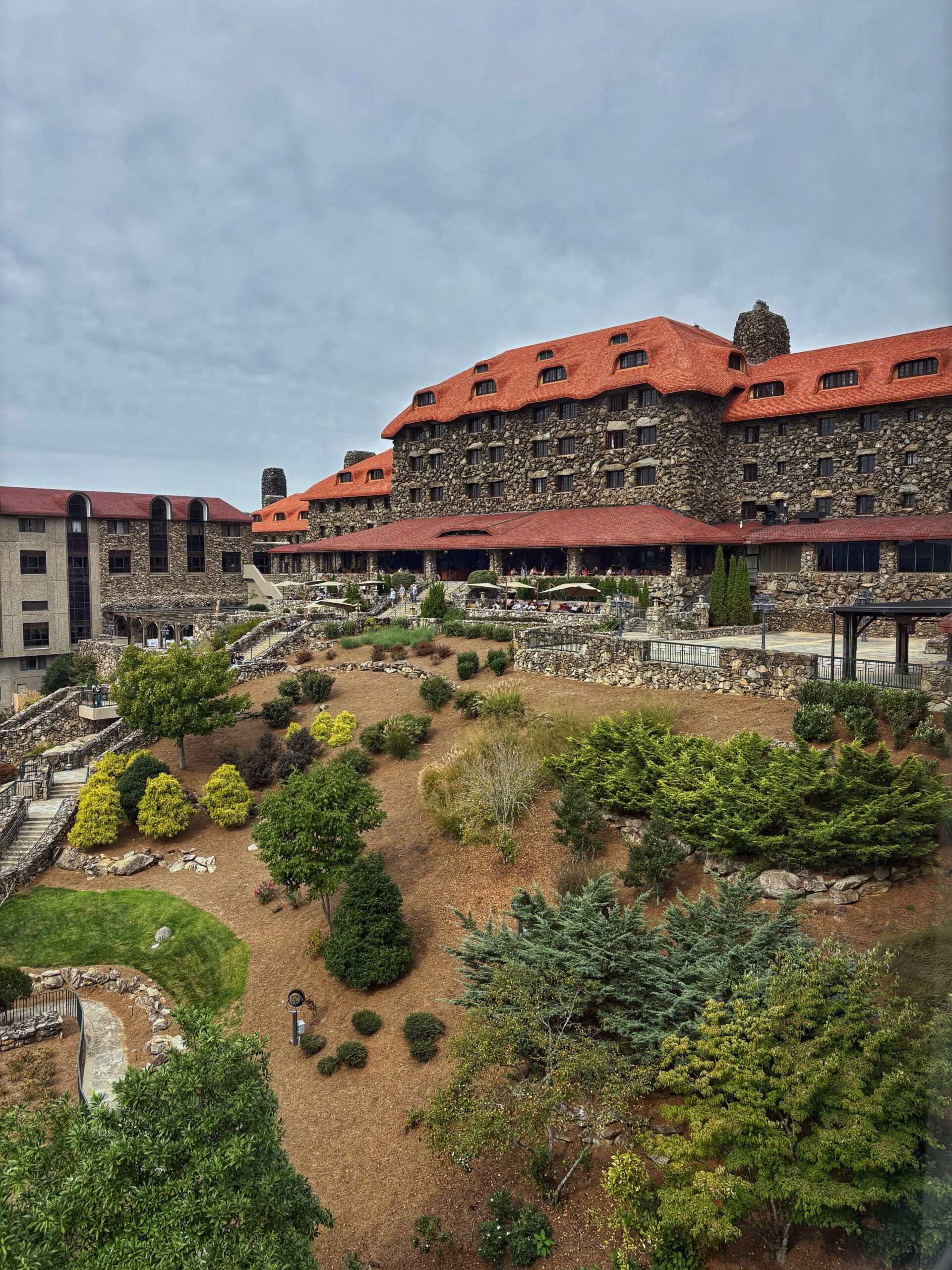 The Omni Grove Park Inn, a large stone building with a red roof, overlooks a tiered hillside garden with shrubs and trees.
